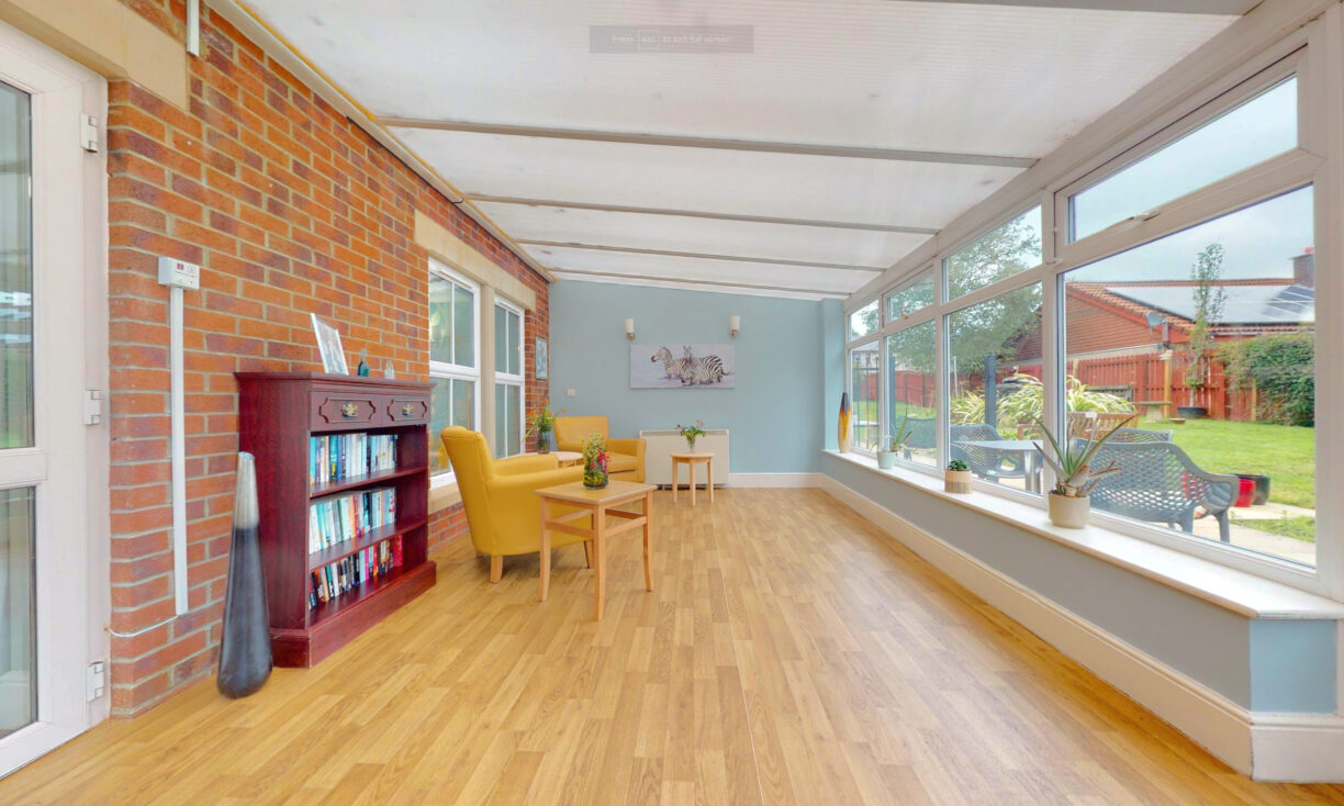A bright sunroom with large windows, a wooden floor, a yellow armchair, a small table, a bookcase, potted plants, and a view of a green garden and red-brick houses outside.