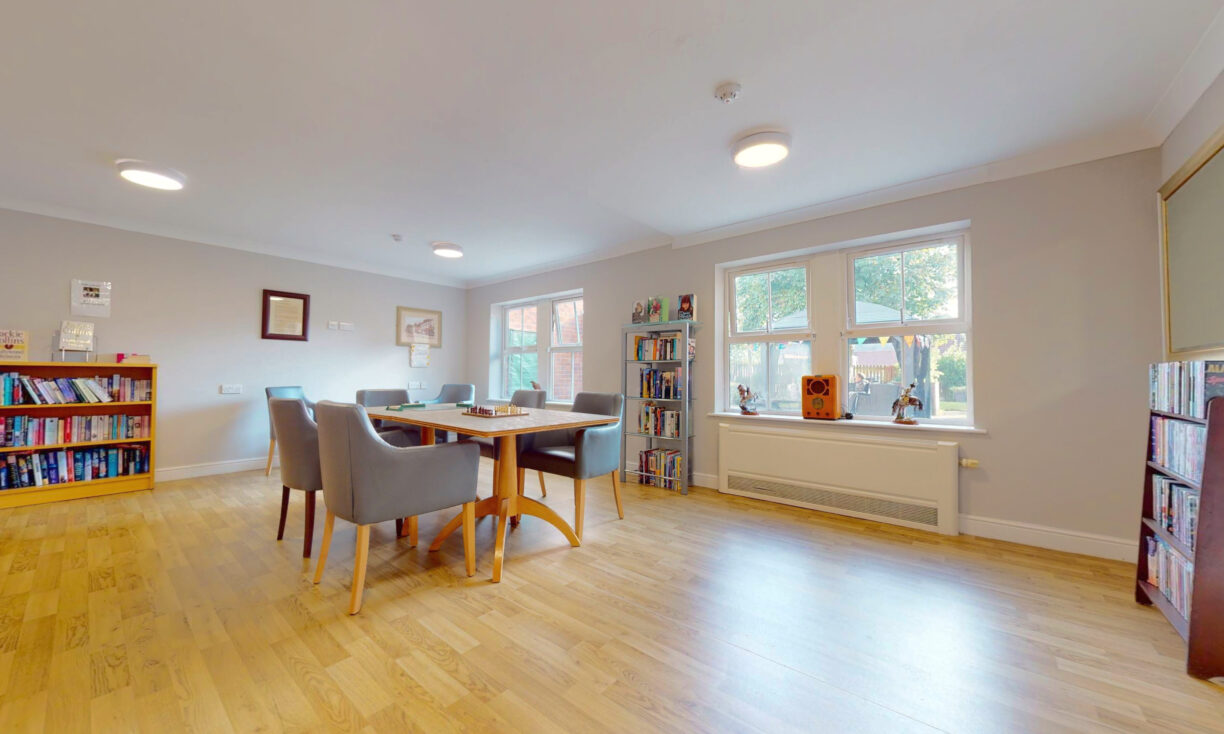 A bright room with wooden flooring, a table and four grey chairs, bookshelves filled with books, and large windows letting in natural light. Art and decor items are displayed on the shelves and windowsill.