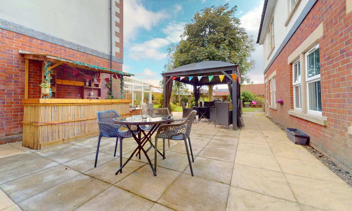 Large paved patio area between brick buildings, featuring a bamboo bar with stools, a round table with chairs, and a gazebo decorated with colourful bunting. A tree and clear sky are visible in the background.