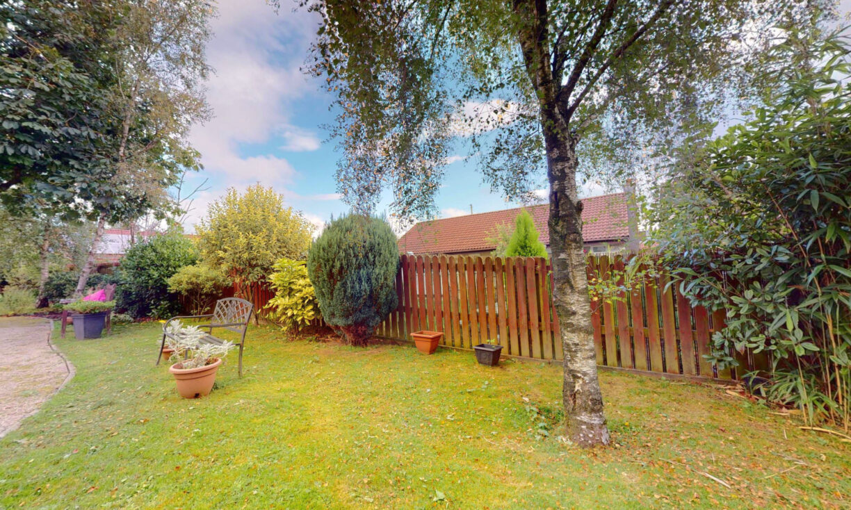A garden with green grass, potted plants, a bench, and a wooden fence. Trees and shrubs border the fence, and a partly cloudy sky is visible overhead.