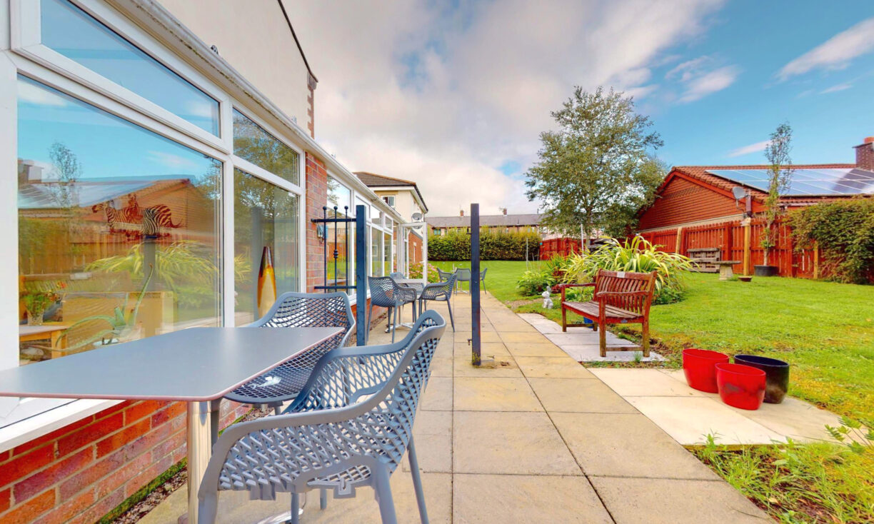 An outdoor patio with tables and chairs beside a building, overlooking a green lawn with benches, plants, and a wooden fence under a partly cloudy sky.