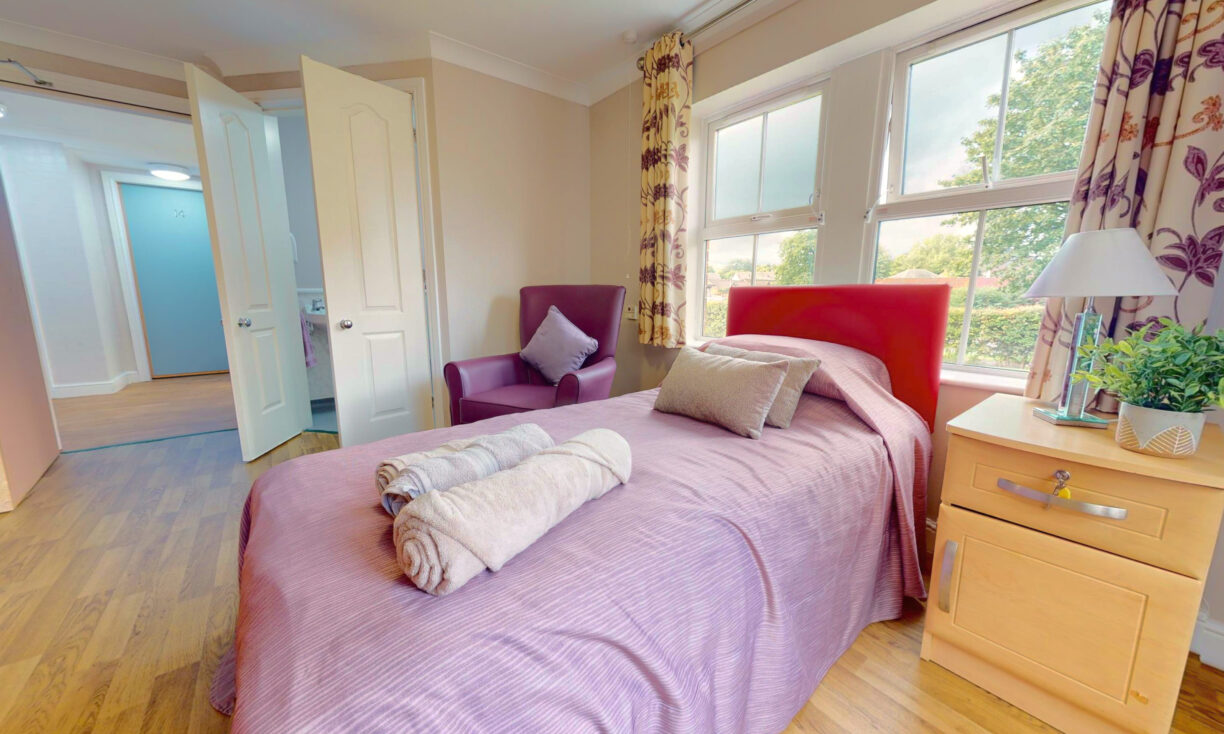 A bright bedroom with a single bed covered in a mauve bedspread, two rolled towels, a purple armchair, a wooden bedside table with a plant, and large windows with floral curtains overlooking trees outside.