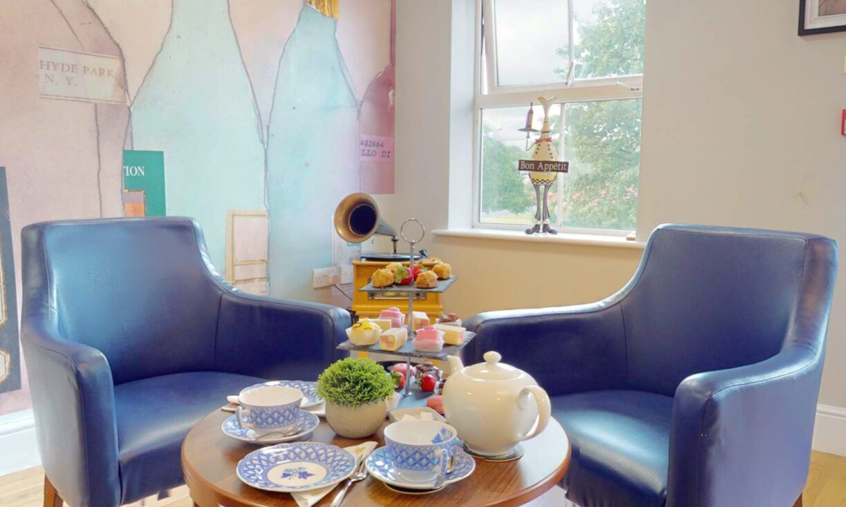 Two blue armchairs face a small round table set for tea with cups, saucers, a teapot, and pastries. A large window and decorative wall art are in the background, creating a cosy, inviting atmosphere.