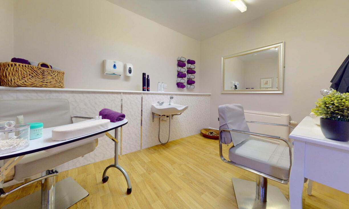 A modern beauty salon room with two chairs, a sink, a desk, neatly folded purple towels, a basket, and a wall mirror, all set on a light wood floor and cream-coloured walls.