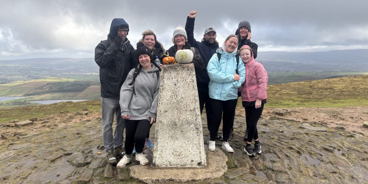 A group of eight people, dressed in warm jackets and rain gear, pose and smile together on a hilltop beside a concrete marker with pumpkins on top. The landscape and cloudy sky stretch out in the background.